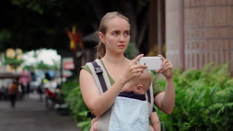 Eine-Junge-Mutter-Mit-Ihrem-Baby-Im-Känguru-Rucksack-Dokumentiert-Ihre-Reise-Durch-Fotos-Auf-Einem-Mobiltelefon.-Während-Sie-Geht,-Wirft-Sie-Einen-Blick-Auf-Den-Telefonbildschirm